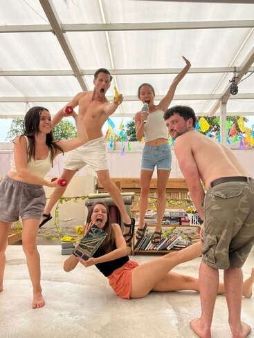 Cinco amigos posam energicamente em um ambiente interno, fazendo posições bobas e criativas. Eles parecem alegres e brincalhões, segurando objetos aleatórios, cercados por decorações brilhantes e uma atmosfera descontraída.