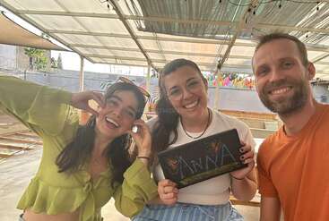 Três pessoas alegres posam juntas em um espaço externo coberto. A mulher no meio segura uma placa decorada que diz "Anna". Elas estão sorrindo e felizes.