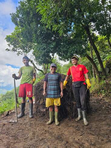 Three men stand smiling outdoors in front of a large pile of organic material, surrounded by lush trees. They wear boots, gloves, and casual clothing, indicating farm work.