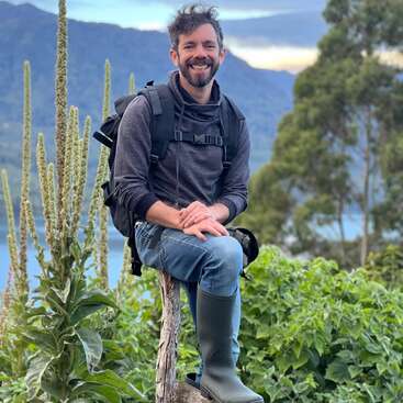 Un homme souriant et barbu, portant un sac à dos, un sweat à capuche, un jean et des bottes en caoutchouc, est assis, heureux, sur une souche d'arbre dans un paysage de montagne pittoresque.