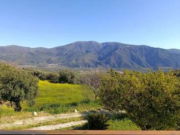 La imagen muestra un paisaje pintoresco con campos verdes, árboles dispersos y una cordillera al fondo, bajo un cielo azul claro en un día soleado.