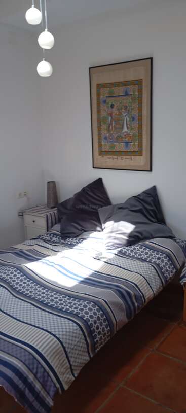 Un dormitorio pequeño y acogedor con ropa de cama a rayas, dos cojines grises, paredes blancas, un cuadro con motivos egipcios, lámparas colgantes y la luz del sol entrando por la ventana.