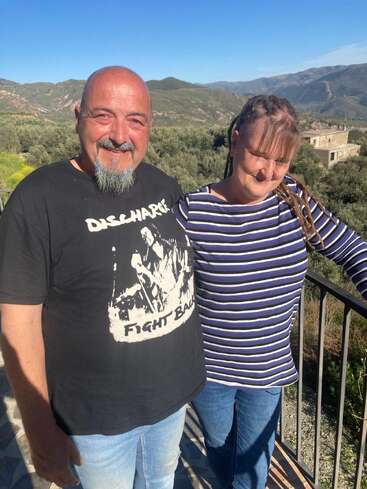 Un hombre y una mujer sonrientes están al aire libre en un día soleado, con colinas verdes y montañas al fondo, disfrutando de la compañía del otro y de las hermosas vistas.