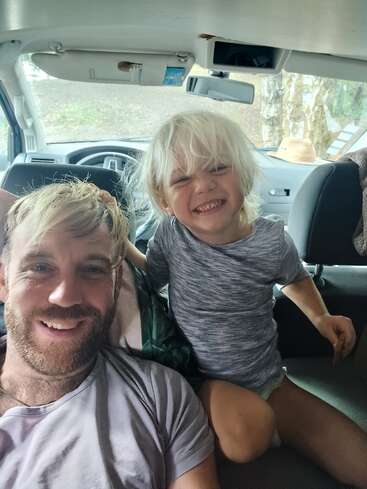 Un homme barbu et un enfant souriant aux cheveux blonds sont assis dans une voiture. Tous deux semblent heureux, profitant d'un moment joyeux ensemble, peut-être pendant un voyage.