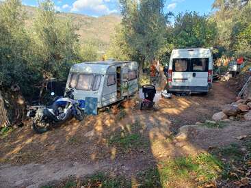 Un petit camping dans une zone boisée comprend une caravane, une camionnette, une moto et une poussette. Des montagnes et des arbres entourent cet espace de vie improvisé.