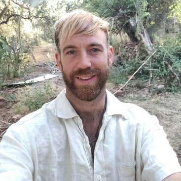 A man with a light beard and blonde hair is smiling, wearing a light shirt, taking a selfie outdoors in a green, wooded area on a sunny day.