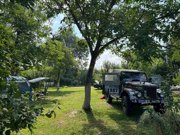 Un clásico todoterreno está aparcado en una zona cubierta de hierba bajo unos árboles, rodeado de tiendas de campaña, sillas de camping y naturaleza, lo que sugiere una tranquila escena de acampada al aire libre.
