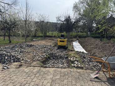 En una obra situada en un jardín se ven una pequeña excavadora amarilla, montones de piedras, una carretilla, árboles en flor y una lona blanca extendida en el suelo.