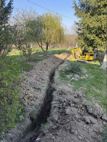 Una miniexcavadora amarilla excava una zanja larga y estrecha en un jardín cubierto de césped y rodeado de árboles y arbustos, probablemente para la instalación de servicios públicos o para trabajos de jardinería.