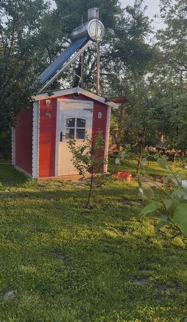 En un frondoso jardín verde se alza un pequeño cobertizo rojo con una puerta blanca. En el tejado del cobertizo hay instalado un calentador de agua solar. Está rodeado de árboles.
