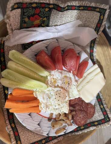 Un plato con huevos fritos, rodajas de tomate, pepino, zanahoria, queso, salami y cebolla descansa sobre un mantel decorativo, acompañado de servilletas en una bandeja de madera.