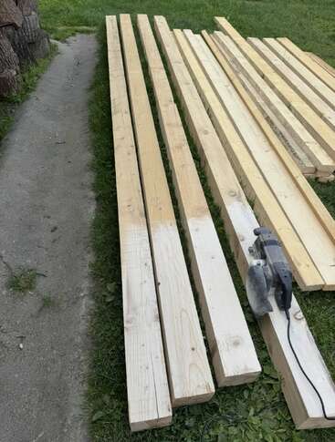 Varias vigas largas de madera están cuidadosamente dispuestas sobre un césped junto a un camino pavimentado, con una sierra circular eléctrica colocada sobre una de las vigas.