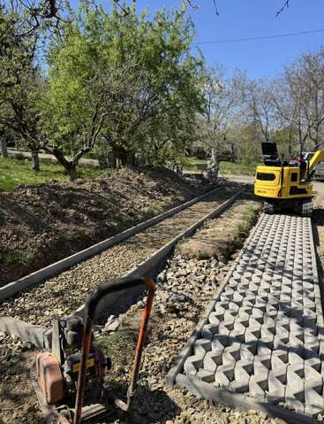 La imagen muestra un camino pavimentado a medio construir en una zona verde y arbolada. Se ven máquinas de construcción, grava y bloques de hormigón que se están colocando para la pasarela.