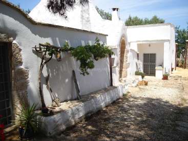L'image représente un bâtiment serein en stuc blanc avec un extérieur en pierre rustique, avec une petite porte voûtée et une cheminée, au milieu d'un environnement extérieur paisible.