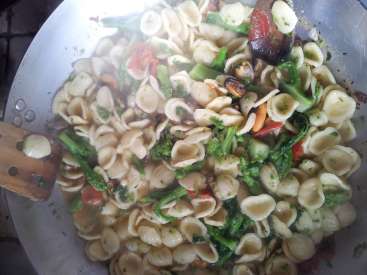 L'image représente une grande casserole en argent remplie d'un plat de pâtes, comprenant des orecchiette, des légumes et une cuillère en bois, probablement en train d'être préparé pour la cuisson.