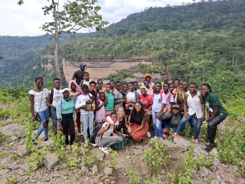 A imagem mostra um grupo de aproximadamente 30 pessoas, a maioria meninas, posando para uma foto em um penhasco rochoso, cercado por uma vegetação exuberante e um cenário montanhoso.