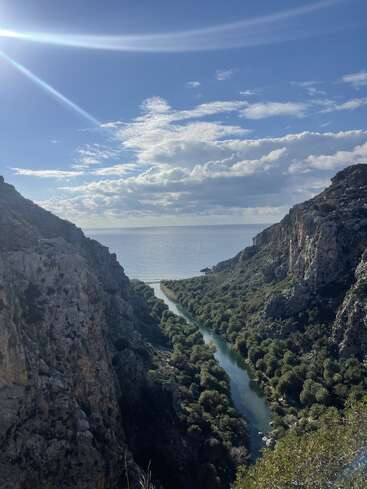 Um rio serpenteia por um vale verdejante e exuberante entre penhascos rochosos, fluindo em direção ao mar cintilante sob um sol forte e um céu salpicado de nuvens.