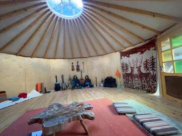 O interior aconchegante de uma yurt com piso de madeira, uma mesa de centro original, instrumentos, almofadas, uma tapeçaria com veados, uma claraboia e duas pessoas sentadas encostadas na parede.