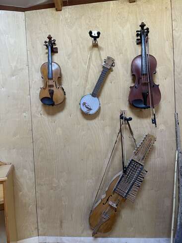 Em uma parede de madeira, dois violinos, um banjo e uma nyckelharpa estão pendurados como decoração, exibindo uma coleção de instrumentos musicais de cordas.