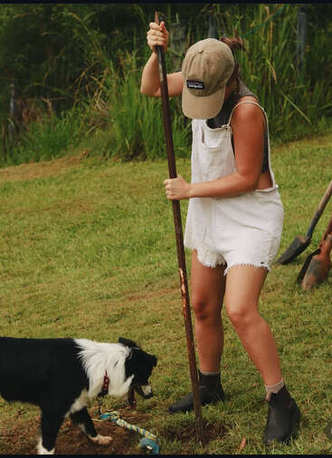 A person wearing white overalls and a cap is digging in grassy ground with a tool, while a black and white dog curiously sniffs nearby.