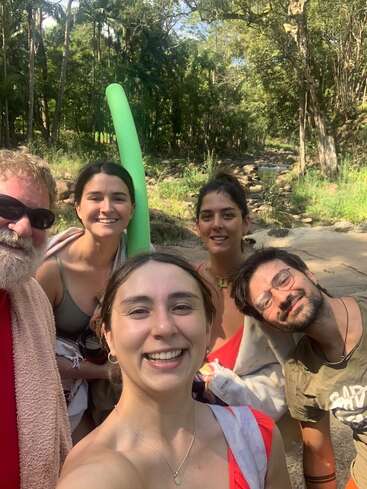 Five people smile for a selfie in a sunny, forested outdoor setting near a rocky stream. They look happy, carrying towels and swim gear, enjoying nature together.