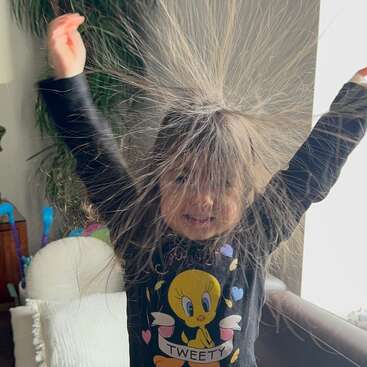 Uma jovem sorridente, com longos cabelos castanhos, está de braços erguidos, com os cabelos soltos devido à eletricidade estática, vestindo uma camiseta preta do Piu-Piu.