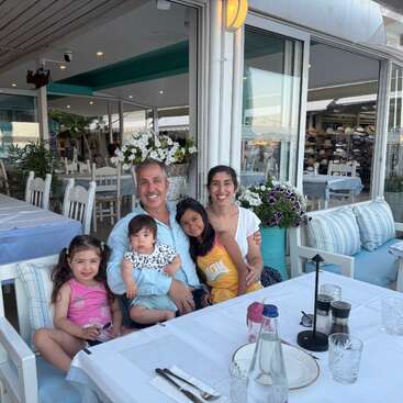Uma família sorridente de cinco pessoas senta-se em uma mesa de restaurante ao ar livre, cercada por flores e decoração aconchegante, desfrutando de um momento feliz e relaxante juntos.