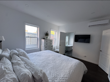 Esta imagem mostra um quarto moderno e bem iluminado com paredes brancas, cama, cômoda, duas janelas, uma TV, ar-condicionado e um banheiro privativo com banheira.