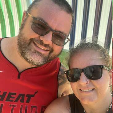 A smiling man and woman pose for a selfie outdoors, both wearing sunglasses. The man has a beard and wears a red sports jersey. Bright background.