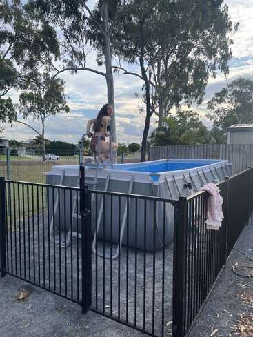 Ein Mädchen im Badeanzug klettert fröhlich in einen oberirdischen Pool, der von einem schwarzen Sicherheitszaun umgeben ist, mit Bäumen und einem bewölkten Himmel im Hintergrund.
