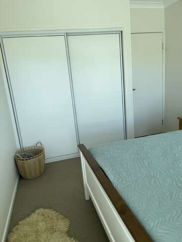 Dieses Bild zeigt eine aufgeräumte Schlafecke mit weißen Wänden, einem doppelten Schiebeschrank, einer weißen Tür, einem Weidenkorb, einem beigen Teppich und einem ordentlich gemachten Bett.