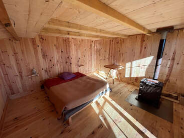 Cette chambre confortable dans une cabane en bois comprend un lit simple avec des couvertures, une petite table d'appoint, un poêle à bois, un éclairage chaleureux et des murs rustiques en bois naturel.