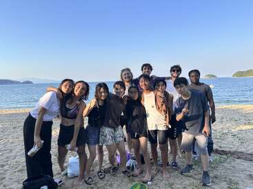 A group of friends pose together on a sandy beach with clear skies and calm water in the background, smiling and enjoying a sunny day by the sea.