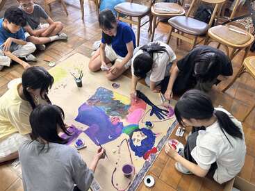 A group of children sit on the floor, collaboratively painting a colorful artwork on a large canvas, surrounded by brushes, paint containers, and wooden chairs.