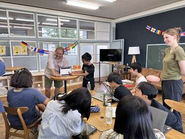 A group of students and two teachers are in a classroom. One student presents something on a laptop while others watch, surrounded by international flags and decorations.