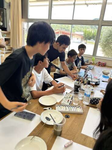A group of students sit around a table with art supplies, watercolors, and brushes, as an adult guides them in a bright, sunlit classroom.