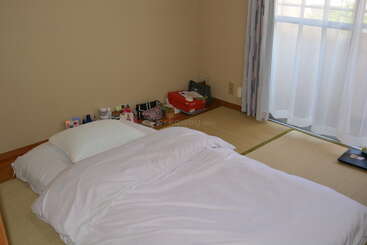 A minimalist room with a futon bed on tatami mats, a white pillow, sunlight through sheer curtains, and neatly organized belongings in the corner. Cozy atmosphere.