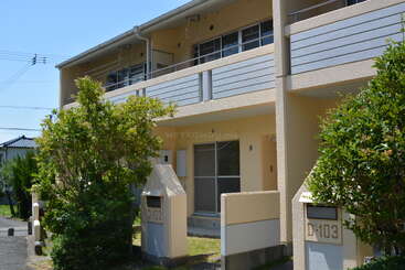This image shows a modern, beige apartment building with balconies, large windows, and greenery. Units D-102 and D-103 are visible, surrounded by bushes.