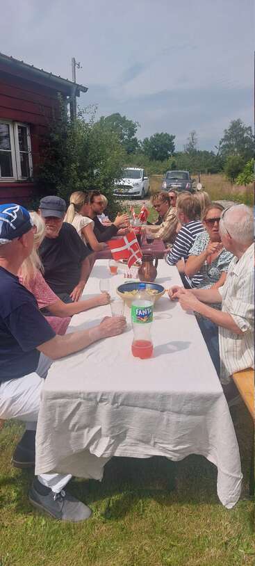 Un grupo de personas está sentado al aire libre en una larga mesa, disfrutando de comida y bebida, rodeados de naturaleza y banderas danesas, en un día soleado.