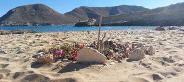 Una escena en una playa de arena con una pequeña disposición de piedras y palos en primer plano, con tranquilas aguas azules y escarpadas montañas bajo un cielo despejado detrás.