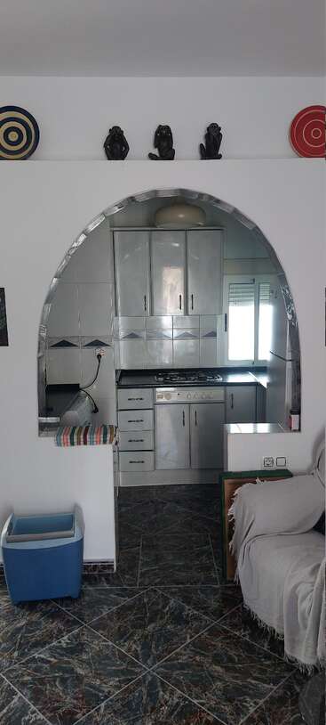 Esta imagen muestra una pequeña cocina con armarios metálicos, vista a través de una abertura arqueada. En la pared superior hay platos decorativos y tres figuras de monos sabios.