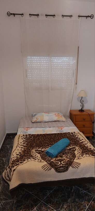 Un dormitorio sencillo con cama individual, manta con estampado de leopardo, toalla azul enrollada, mesilla de noche de madera, lámpara, sábanas de temática náutica y cortinas blancas detrás de una ventana cerrada.