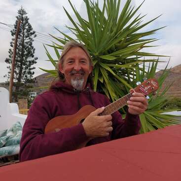 Un hombre sonriente con pelo largo y barba toca el ukelele al aire libre. Lleva una sudadera con capucha granate y está sentado frente a una planta alta de color verde con pinchos.