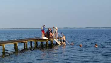 Several people gather on a wooden pier overlooking calm blue water. Some sit, relax, or converse, while others swim and play in the inviting sea below.