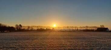 The image shows a tranquil sunrise over a frosty field, with silhouettes of trees and a few houses lining the horizon under a clear sky.