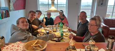 A group of seven people sits around a wooden table, enjoying a meal together indoors. They are smiling, giving thumbs up, and appear happy and relaxed.