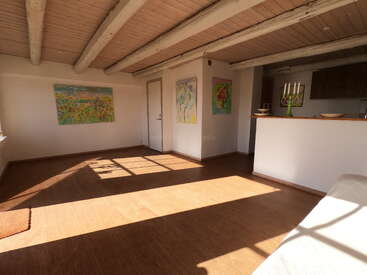 Sunlit room with wooden ceiling beams, light walls, and brown flooring. Colorful abstract paintings hang on the walls. Kitchen counter, candlestick, and ceramics visible. Cozy atmosphere.