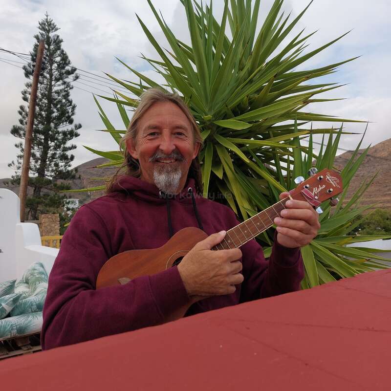 Un hombre sonriente con pelo largo y barba toca el ukelele al aire libre. Lleva una sudadera con capucha granate y está sentado frente a una planta alta de color verde con pinchos.