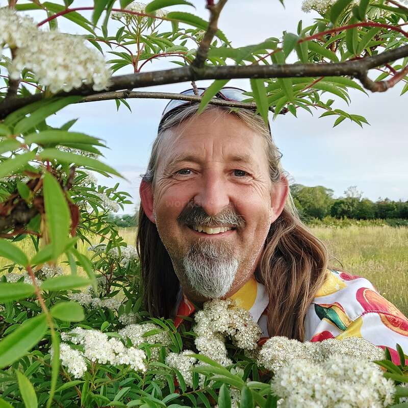 A smiling man with long hair and a colorful shirt stands among white flowering bushes, outdoors in a green field under a partly cloudy blue sky.