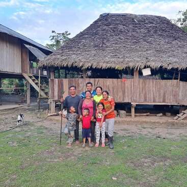 Un groupe de personnes souriantes, adultes et enfants, se tient devant une maison rustique en bois au toit de chaume, dans un village.